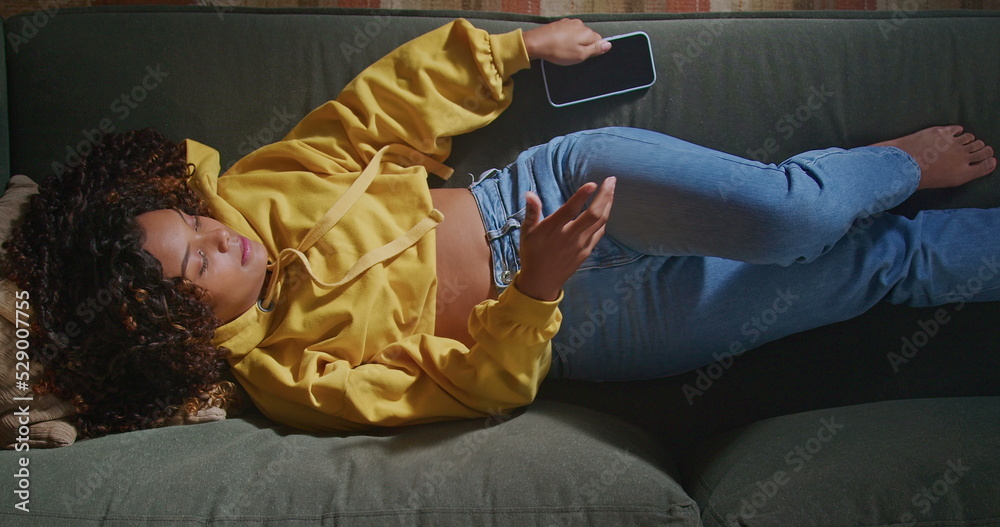 One hispanic black girl putting cellphone away. From above angle of an African American young woman resting on sofa. Restful person relaxing in the evening at living room home napping