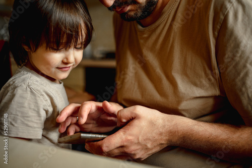 The father sits at home at the table and looks at the mobile phone, next to him stands a little son and peeps into the screen of the phone. The use of gadgets in modern life