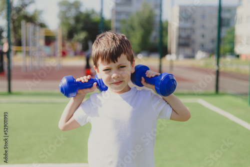 A child with dumbbells on the playground. A sporty kid. Illustrating an article about sports. Children's exercises. Education of champions. Light weightlifting.