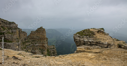 Dramatic landscape - panoramic view of a hilly green valley blurred in a misty haze and steep stone cliffs from the Bermamyt plateau in Karachay-Cherkessia on a cloudy summer day