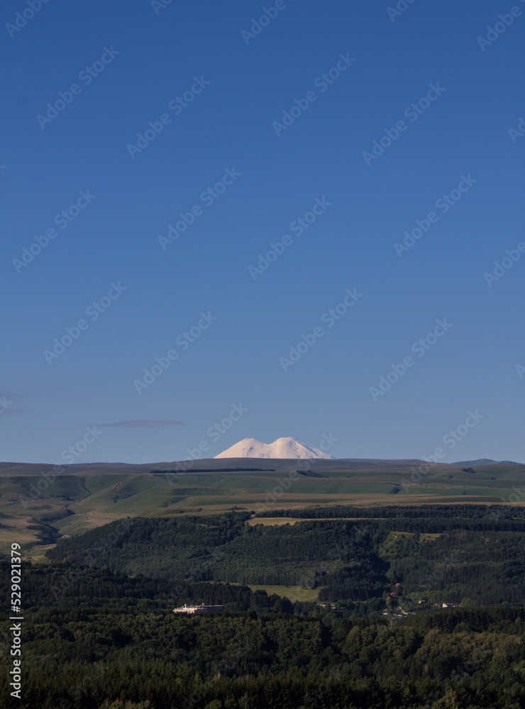 Panoramic view of the picturesque green hilly valley and the snowy peak of Mount Elbrus blurred in a misty haze on a clear sunny summer day