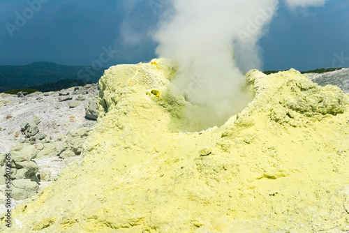 cone of sulfur deposits around a fumarole in a solfataric field illuminated by the sun against a stormy landscape in the distance photo