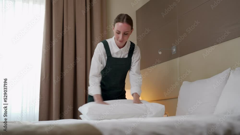 Female employee makes bed changing fresh linen and folding white pillows. Young maid in uniform takes care of order and cleanliness in hotel room