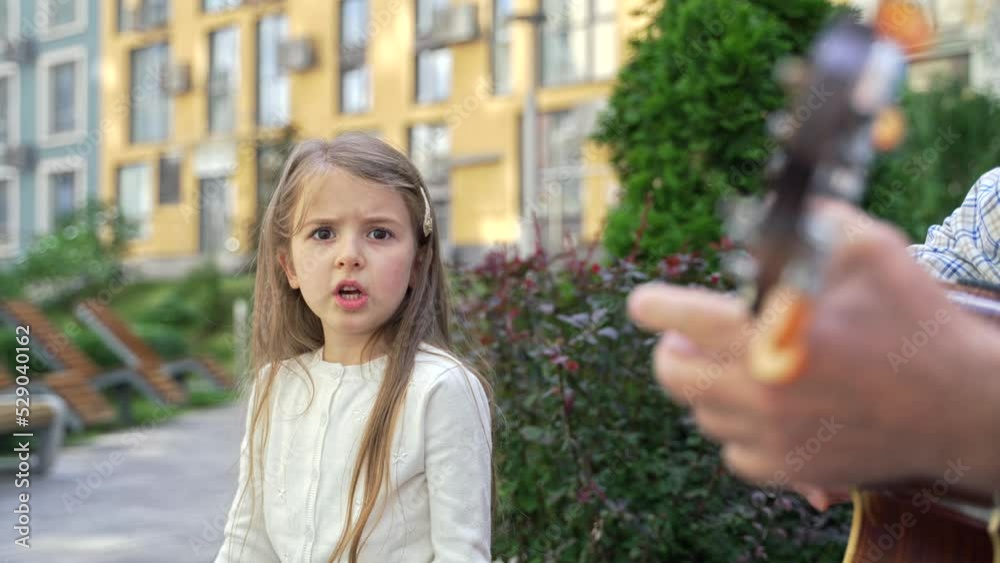Close up of serious pretty little girl singing piercingly while looking at the camera outdoor. Cute child girl with long blonde hair singing to the accompaniment of a guitar sitting on the bench Close up of serious pretty little girl singing piercingly while looking at the camera outdoor. Cute child girl with long blonde hair singing to the accompaniment of a guitar sitting on the bench