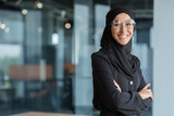 Portrait of muslim woman in hijab at work in office, business woman smiling and looking at camera with crossed arms.