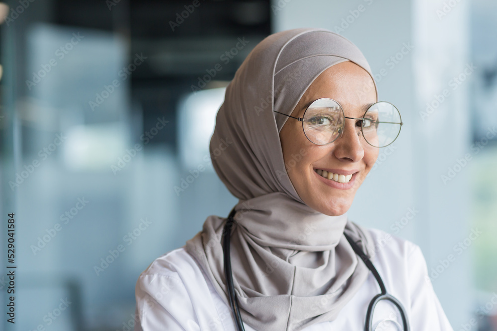 Close-up photo portrait of Muslim female doctor in gray hijab, female doctor smiling and looking at camera, female worker in glasses and white medical coat and glasses working inside modern clinic