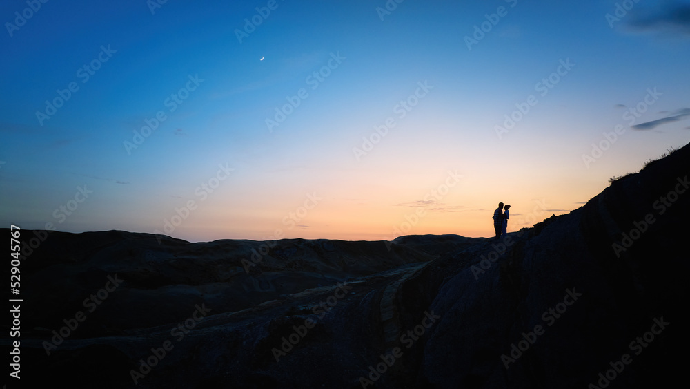 Silhouette of a couple of young people embracing on the edge of a cliff against a colourful evening sky. Love and relationships. 