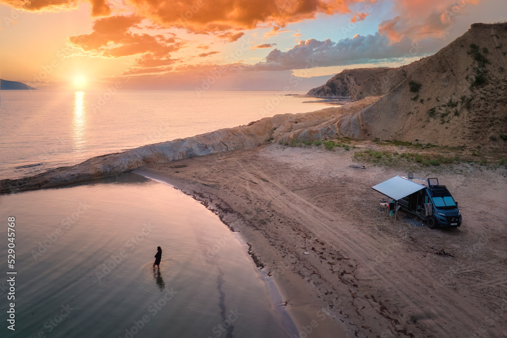 Aerial photo of campervan on abandoned beach against beutiful sunset. People bathing in the sparkling sea. Outdoor nomad lifestyle, van life holiday. Independent road trip concept. - obrazy, fototapety, plakaty 