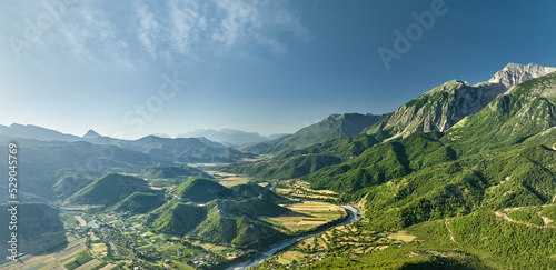 Aerial panoramic view of the Vjose River valley in Albania. The crystal clear, turquoise river winds its way through fields against a backdrop of majestic mountains. Blue skies, summer.  photo