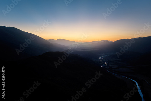 Aerial panoramic view of the dark valley of the Vjose River after sunset, surrounded by the silhouettes of majestic mountains. Orange-blue sky, sun rays behind the mountains, summer.  photo