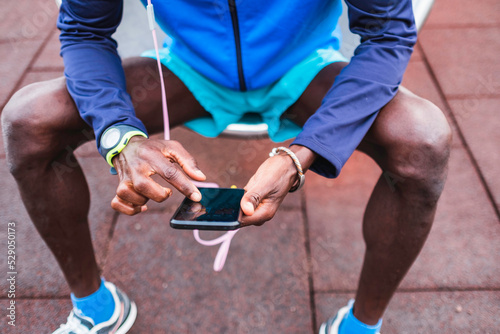 black man sitting in a playground in sportswear, listening to music with headphones through his mobile phone. 