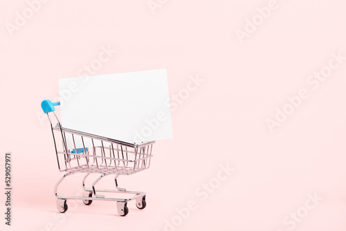 Shopping cart with a blank white paper card on a pastel pink background. Minimalist design with copy space. Concepts: market deals, seasonal sales and discounts, black friday.