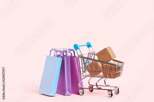 Shopping cart with cardboard boxes and paper bags on a pastel pink background. Simple design with copy space. Concepts: market sales, seasonal discounts, black friday, logistics, packaging.