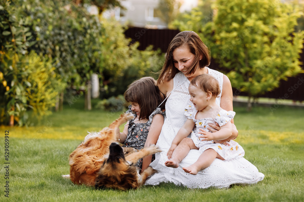 Fototapeta premium Family day, mother's day. Beautiful smiling young mom and two child daughters cuddling happy domestic dog on the backyard lawn. Idyllic family having fun with pet outdoors on summer holiday.