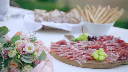 Exclusive banquet table with variety of meat cut on handmade wooden board decorated with fresh grapes, bowl with bread sticks and slices, peak of white clean plates prepared for invited guests