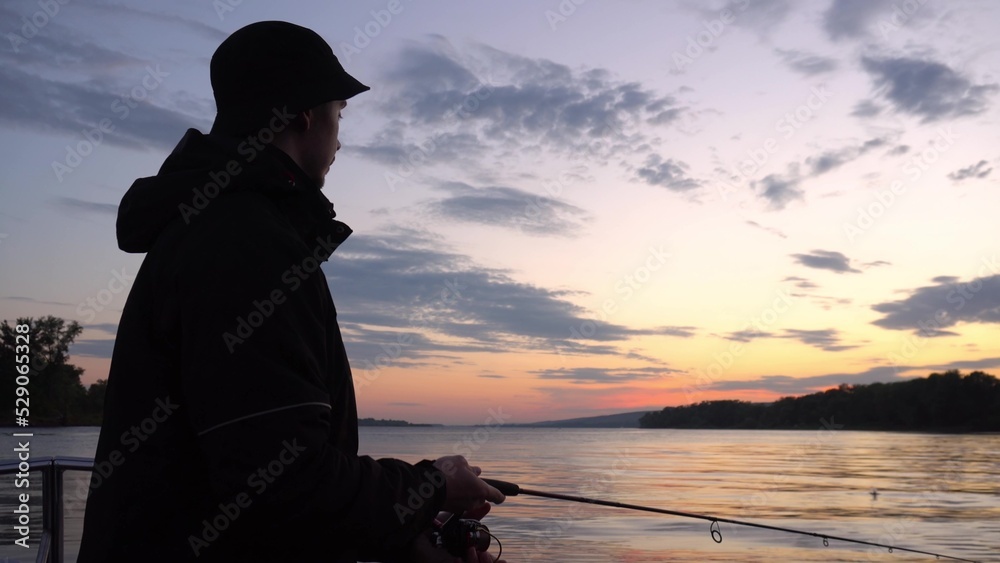 Fototapeta premium A fisherman in a jacket is fishing at sunset on the river from a boat. A young man throws a bait on a fishing rod and spins the spinning.