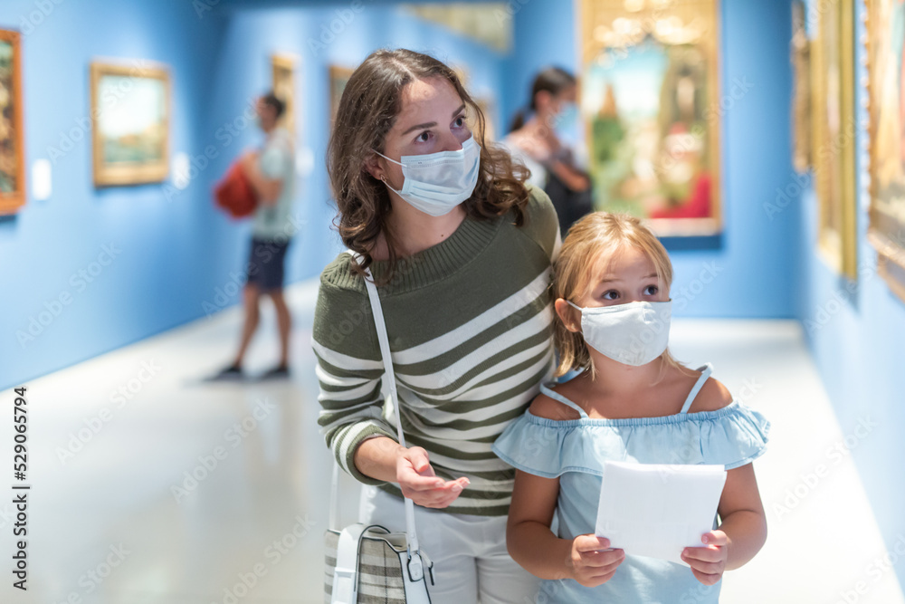 Mother and daughter in protective medical masks looking at paintings in halls of museum