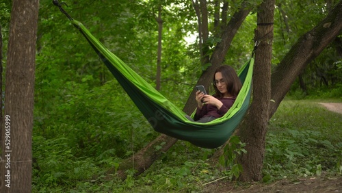 A young woman lies in a hammock in the woods with a phone in her hands. A girl with glasses chats on the Internet through a smartphone. The camera is getting closer.
