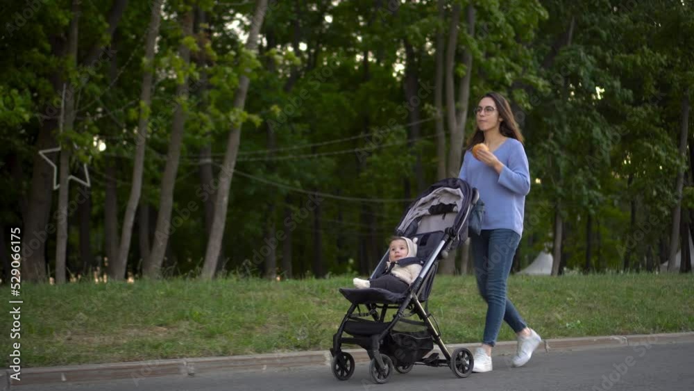 Mom walks with a baby in a stroller and eats a burger on the go. A young woman in glasses pushes a stroller with her son and eats a burger in the park. Mom walks with a baby in a stroller and eats a burger on the go. A young woman in glasses pushes a stroller with her son and eats a burger in the park.