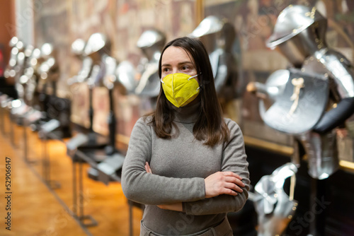 Interested young woman wearing yellow protective face mask viewing collection of medieval knights armor in historical museum. Forced precautions in pandemic © JackF