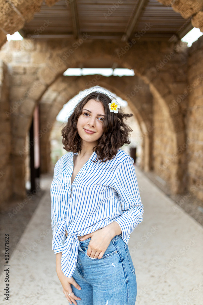 A pensive brunette girl with curly long hair in a white linen shirt with a blue stripe, blue jeans and green sneakers with white plumeria flower in setting sun against the backdrop of medieval city. 