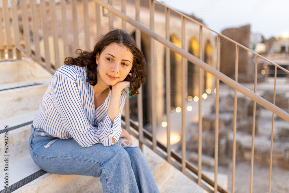 A pensive brunette girl with curly long hair in a white linen shirt with a blue stripe, blue jeans with a white plumeria flower in the setting sun against the backdrop of a ruined medieval city. 