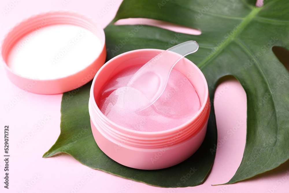 Under eye patches in jar with spatula and green leaf on light pink background. Cosmetic product Under eye patches in jar with spatula and green leaf on light pink background. Cosmetic product