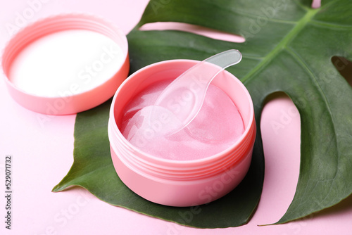 Under eye patches in jar with spatula and green leaf on light pink background. Cosmetic product