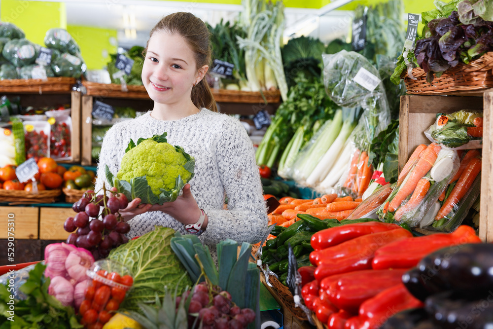Portrait of happy little girl standing among fresh fruits and vegetables on store shelves