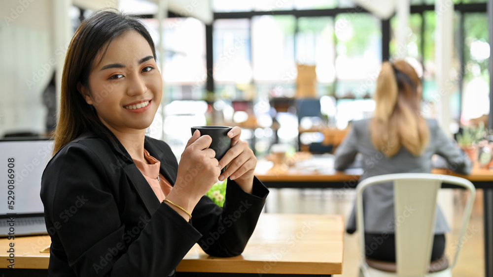 Asian businesswoman at the coffee shop, holding a coffee cup and smiling to camera.