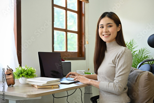 Canvastavla Attractive Asian businesswoman sitting at her office desk, smiling and looking at camera