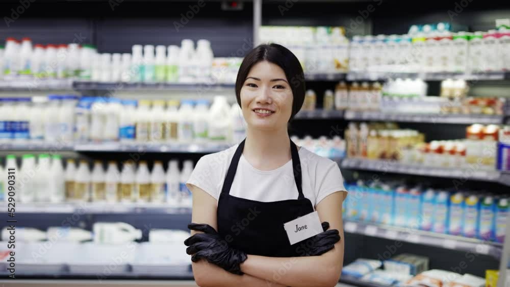 Portrait of an attractive young Asian saleswoman in a black apron standing in a supermarket with shelves of dairy products in the background, looking at the camera and smiling. Trade business and Portrait of an attractive young Asian saleswoman in a black apron standing in a supermarket with shelves of dairy products in the background, looking at the camera and smiling. Trade business and