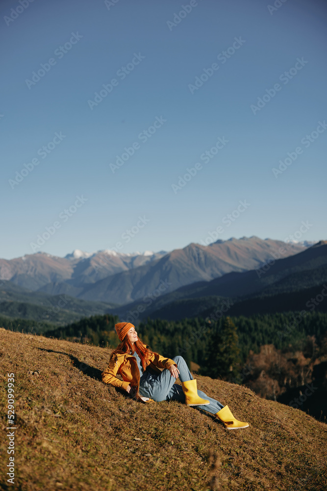 Fototapeta premium Woman full-length sitting resting on a hill and looking at the mountains in a yellow raincoat and jeans happy camping trip in the winter, freedom lifestyle 