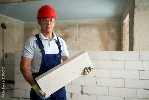 Bricklayer or mason constructs wall of autoclaved aerated concrete blocks. Portrait of brickwork worker contractor standing with quality foamed concrete doing masonry on construction site.