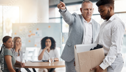 Black man, sad and fired by boss in a meeting holding a box in disappointment at the office. Manager or company leader pointing to the exit and firing employee in front of colleagues at the workplace