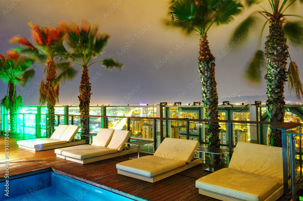 Fototapeta premium Patio lounge chairs at night next to a pool overlooking a city.