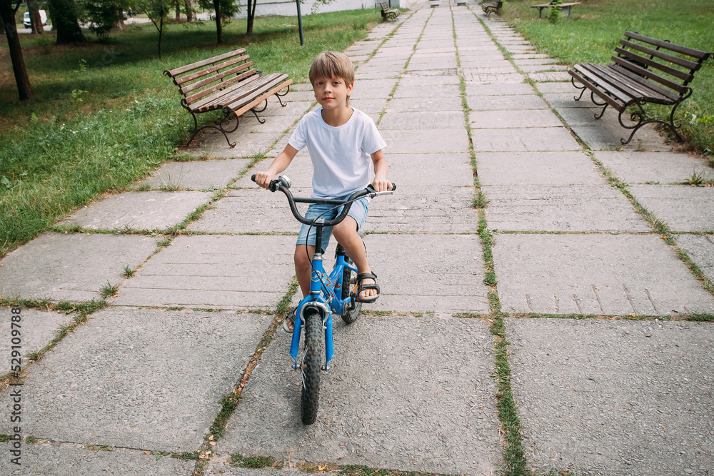 Obraz premium A little cute boy in denim shorts and a white t-shirt rides a children's bike along a path in the park during the day.