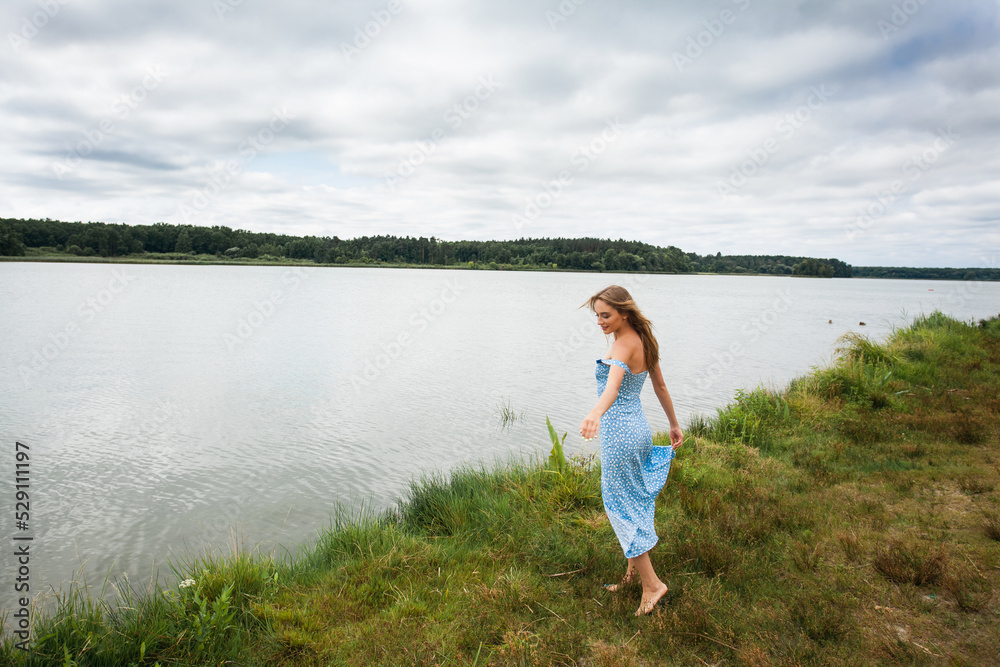 A beautiful slender smiling girl with long blond hair, in a blue summer dress, spins and walks along the river bank, against the backdrop of a picturesque landscape. A beautiful slender smiling girl with long blond hair, in a blue summer dress, spins and walks along the river bank, against the backdrop of a picturesque landscape.