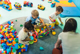 Toddlers and their nursery teacher playing with plastic building blocks and colorful car toys in a nursery school playroom. Early brain and skills development. High quality photo