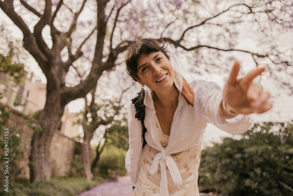 Smiling young caucasian woman reaches for camera, enjoys time in nature. Brunette wears sundress and shirt in summer. Relaxed lifestyle, concept Smiling young caucasian woman reaches for camera, enjoys time in nature. Brunette wears sundress and shirt in summer. Relaxed lifestyle, concept