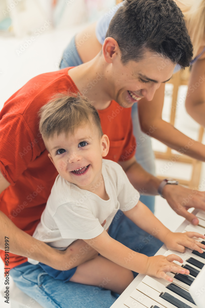 Fun childhood concept. Music in the life of young children. Happy smiling proud caucasian dark-haired dad playing the piano with his smiling at camera son. Father and son relationship. High quality Fun childhood concept. Music in the life of young children. Happy smiling proud caucasian dark-haired dad playing the piano with his smiling at camera son. Father and son relationship. High quality