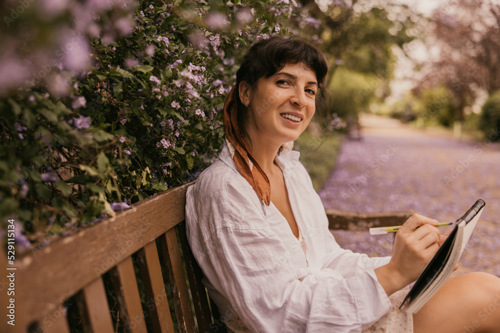 Beautiful caucasian young woman smiling looking at camera, sitting on bench and drawing in album. Brunette wears sundress and shirt outdoors. Inspiration concept Beautiful caucasian young woman smiling looking at camera, sitting on bench and drawing in album. Brunette wears sundress and shirt outdoors. Inspiration concept