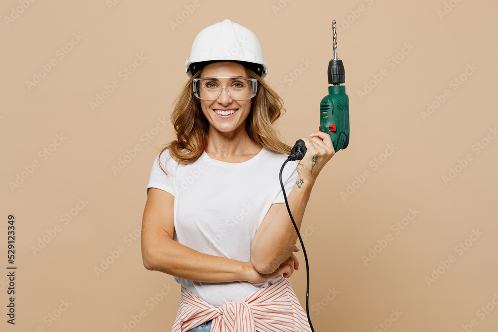Young smiling confident employee laborer handyman woman in white t-shirt helmet glasses hold drill isolated on plain beige background Instruments accessories for renovation room Repair home concept. Young smiling confident employee laborer handyman woman in white t-shirt helmet glasses hold drill isolated on plain beige background Instruments accessories for renovation room Repair home concept.