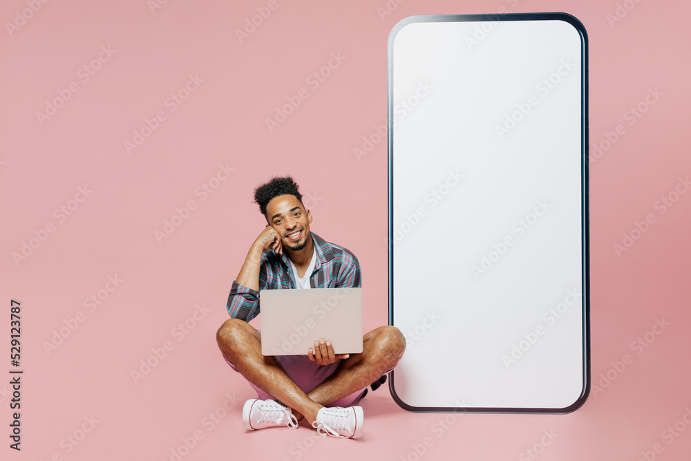 Full body young man of African American ethnicity in blue shirt sit near big huge blank screen mobile cell phone copy space mockup area laptop computer isolated on plain pastel light pink background.