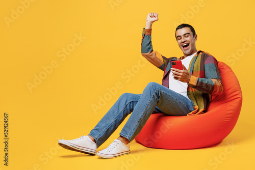 Fotografija Full body young overjoyed happy fun middle eastern man he wearing casual shirt white t-shirt sit in bag chair hold in hand use mobile cell phone do winner gesture isolated on plain yellow background