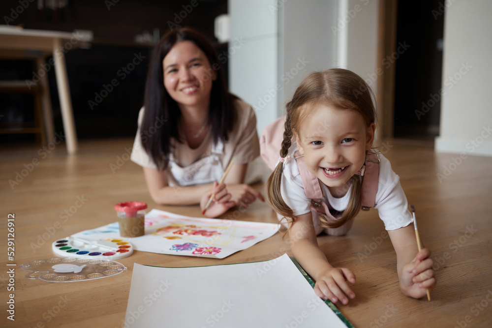 Smiling young mother and little daughter lie on a warm wooden floor in the living room, drawing together, happy mother rejoices at her daughter's drawings. paint on canvas Smiling young mother and little daughter lie on a warm wooden floor in the living room, drawing together, happy mother rejoices at her daughter's drawings. paint on canvas