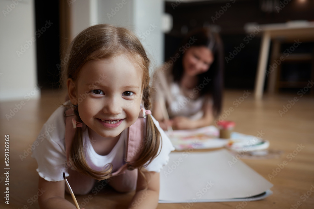 Smiling young mother and little daughter lie on a warm wooden floor in the living room, drawing together, happy mother rejoices at her daughter's drawings. paint on canvas Smiling young mother and little daughter lie on a warm wooden floor in the living room, drawing together, happy mother rejoices at her daughter's drawings. paint on canvas