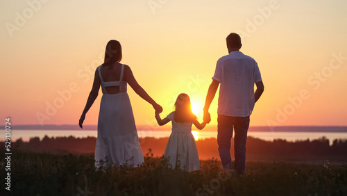 Happy parents silhouettes with excited daughter walk on evening meadow. Family looks together at bright sunset walking on lawn against river