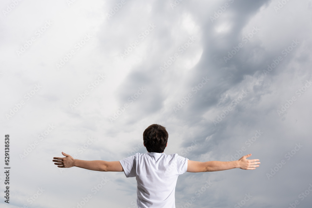 Rear view of boy with open arms breathing and looking at dramatic stormy sky. Praying for rain concept Rear view of boy with open arms breathing and looking at dramatic stormy sky. Praying for rain concept