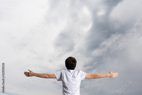 Rear view of boy with open arms breathing and looking at dramatic stormy sky. Praying for rain concept